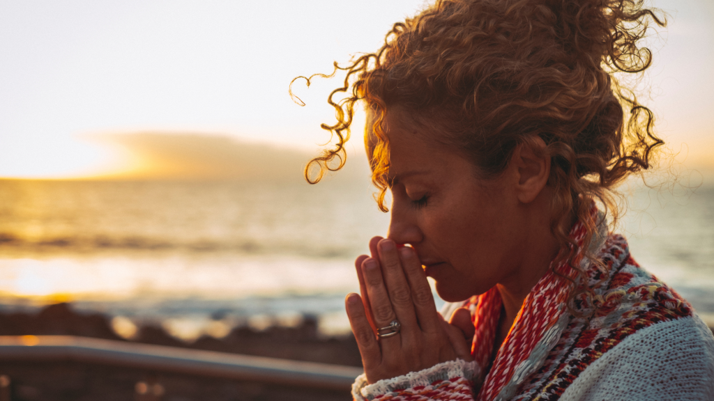 woman on a beach- eyes closed with hands in prayer position near face.